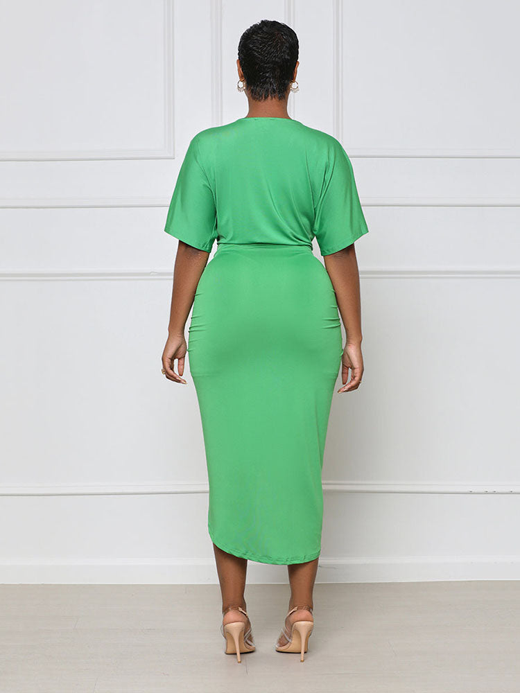 Back view of a woman wearing a green V-neck, short-sleeve, ruched, draped midi dress with a tulip hem.