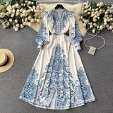 A white maxi dress with a blue paisley print laid flat on a grey surface, adorned with floral arrangements and accessories.