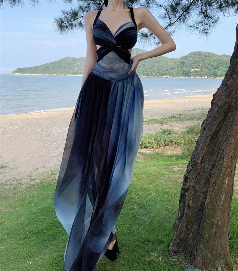 Full view of a woman in a dark blue and black ombre dress with a high slit, standing outdoors by the water.