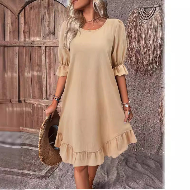 Woman wearing a tan short puff sleeve A-line dress with a ruffled hem and neckline, holding a straw hat.