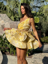 Woman wearing a yellow floral print one-shoulder mini dress with a tiered ruffled skirt, posing outdoors.