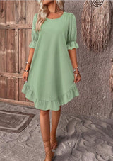 Woman wearing a light green short puff sleeve A-line dress with a ruffled hem and neckline, holding a straw hat.