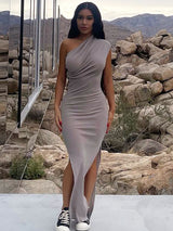 Woman wearing a grey one-shoulder ruched maxi dress with a high slit and sneakers, standing outdoors with a rocky, mountainous background.