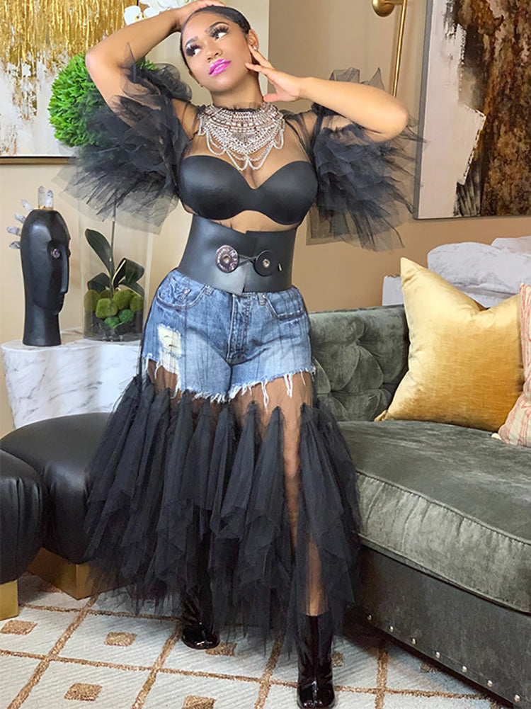 Woman in a black sheer mesh maxi dress with voluminous ruffle sleeves and hem, styled with denim shorts and a black leather belt.