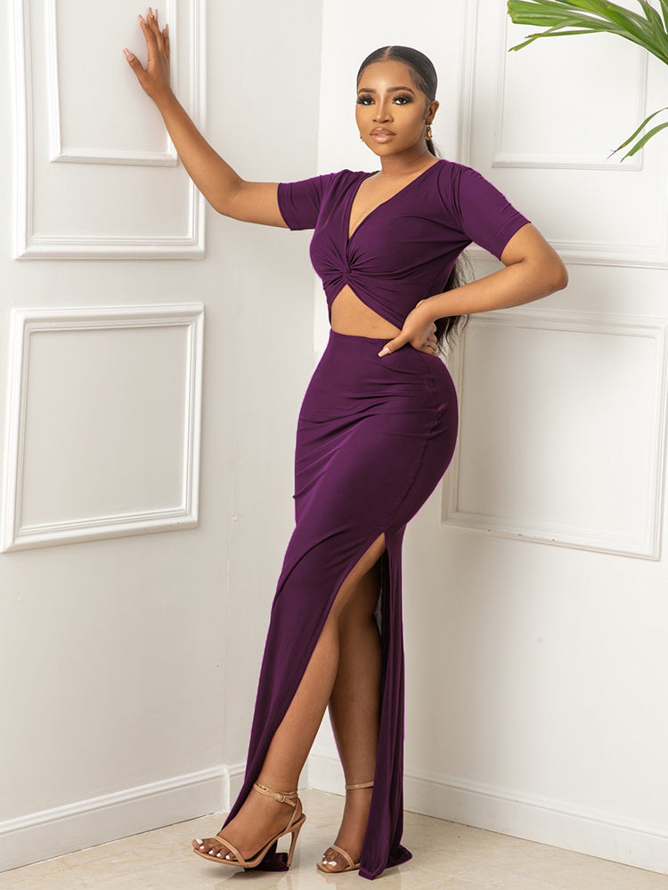 Woman wearing a purple two-piece set with a twisted v-neck top and a high slit skirt, posing with her hand on a white wall.