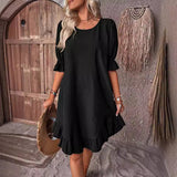 Woman wearing a black short puff sleeve A-line dress with a ruffled hem and neckline, holding a straw hat.