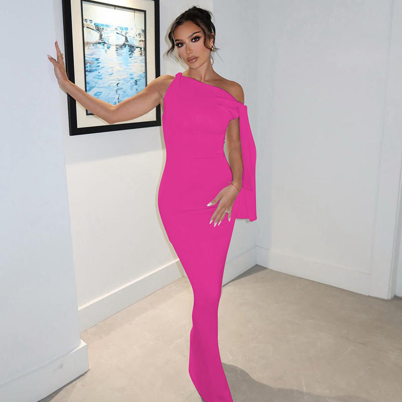 Woman in a bright pink floor-length dress with an asymmetrical neckline and draped shoulder detail, leaning against a white wall.
