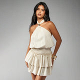 Woman wearing a cream halter neck mini dress with a smocked waist and tiered skirt, looking at the camera.