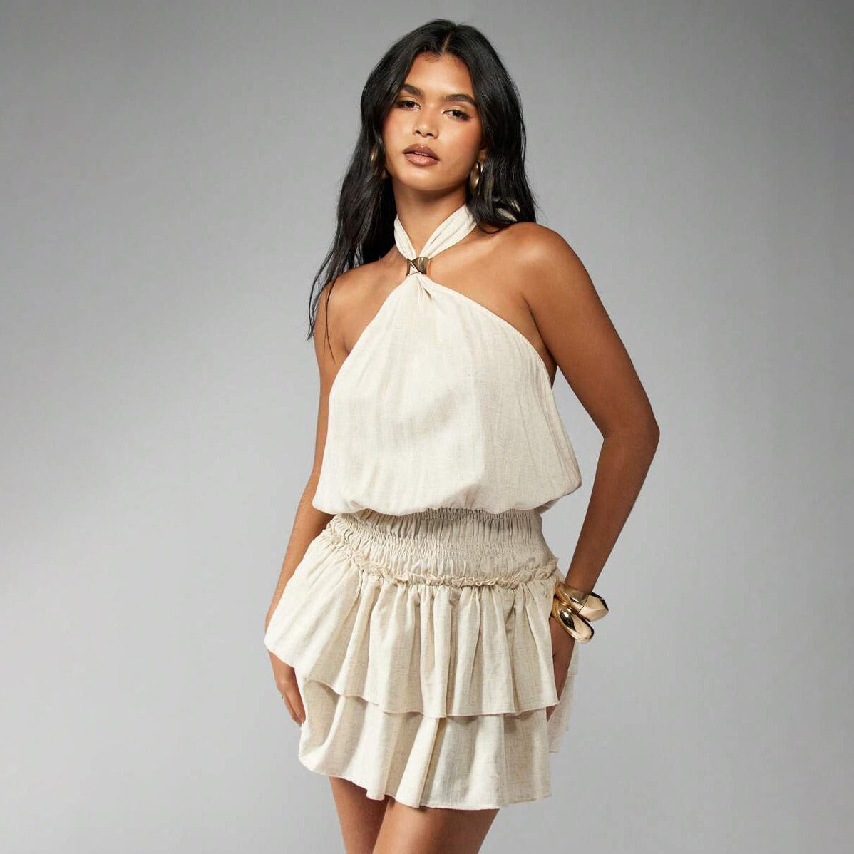 Woman wearing a cream halter neck mini dress with a smocked waist and tiered skirt, looking at the camera.
