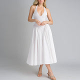 Woman wearing a white halter neck midi dress with a deep V-neckline and full skirt, posing with hands clasped, against a gray background.