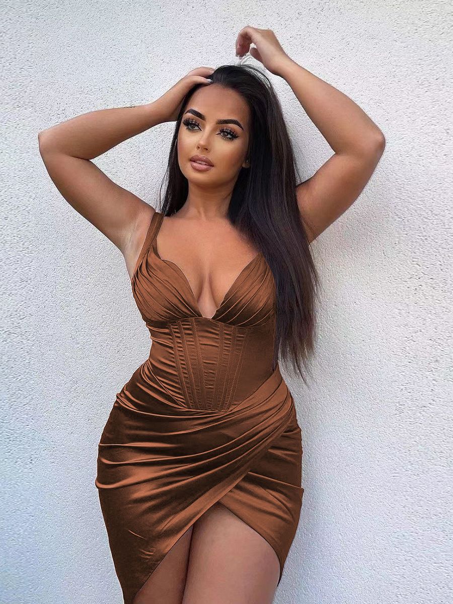 Woman with dark hair wearing a brown satin dress with a plunging neckline and high slit, posing against a textured wall.
