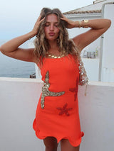 Woman wearing an orange mini dress with starfish embellishments and ruffled hem, gold jewelry, and a beaded bag, posing outdoors with the ocean in the background.