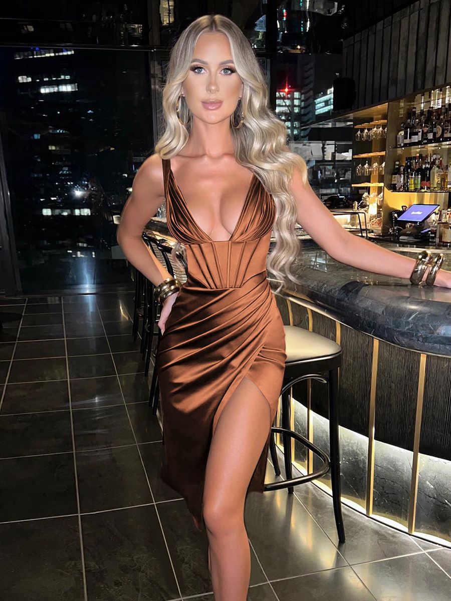 Woman in a brown satin dress with a plunging neckline and high slit standing by a bar.