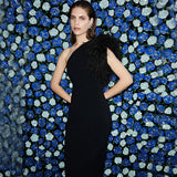 Woman in a black one-shoulder gown with feather accent posing against a blue and white floral backdrop.