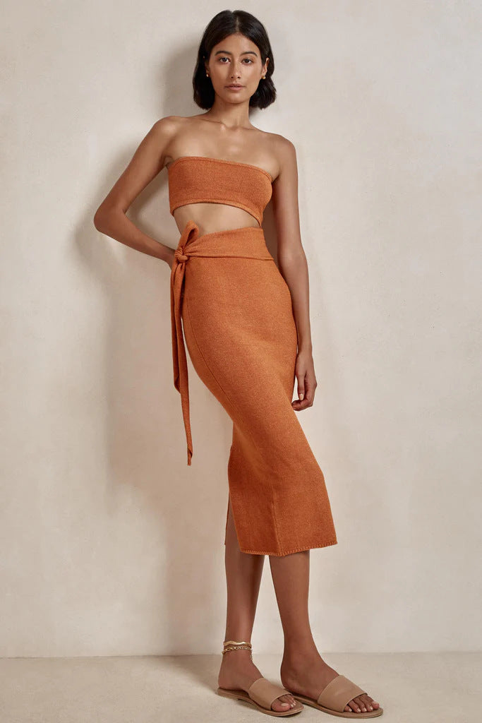 Woman wearing an orange strapless knit crop top and matching midi skirt with a tie waist and side slit, shown from the front.