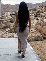 Rear view of a woman walking away, wearing a grey one-shoulder ruched maxi dress with a high slit and sneakers, outdoors.