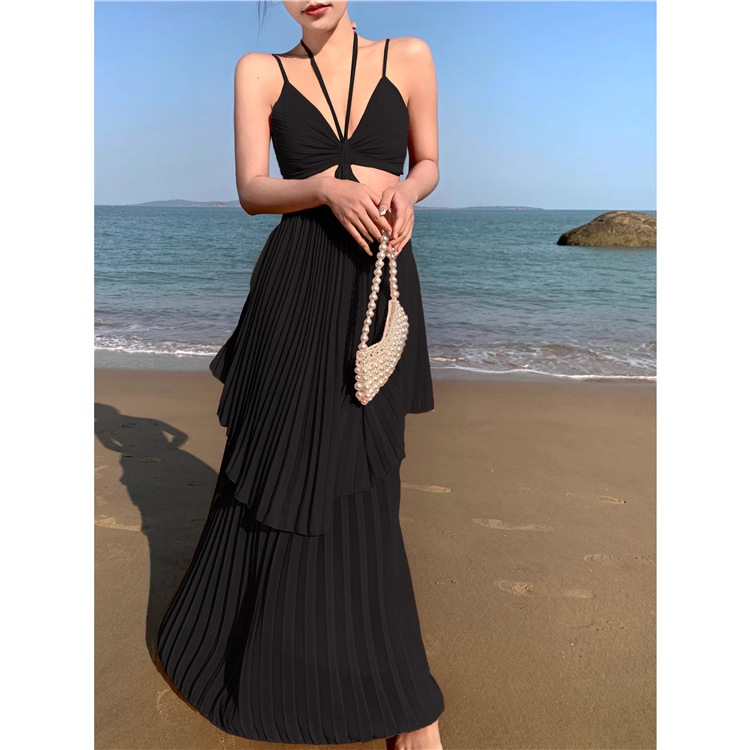 Woman wearing a black halter neck maxi dress with a cut-out and pleated tiered skirt, holding a beaded clutch on a beach.