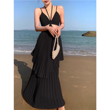 Woman wearing a black halter neck maxi dress with a cut-out and pleated tiered skirt, holding a beaded clutch on a beach.