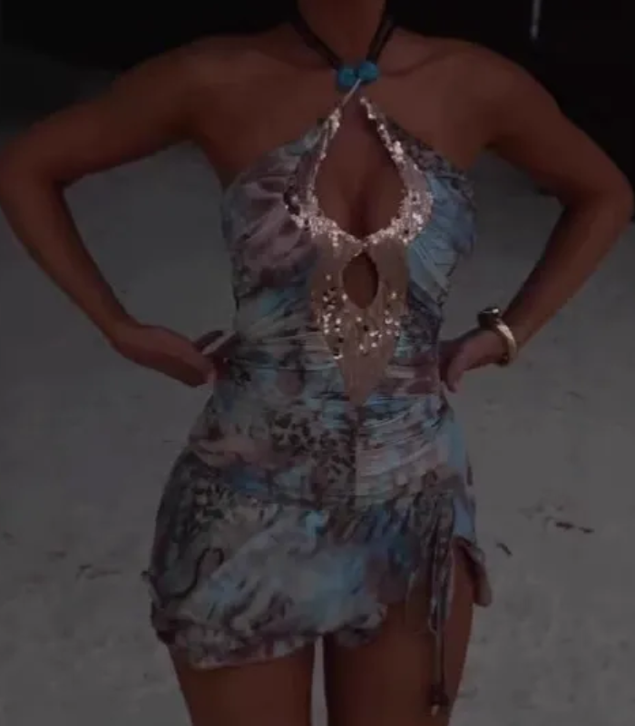 Woman standing in a light blue and brown abstract print halter neck mini dress with a sequined keyhole neckline and turquoise necklace, with hands on her hips.