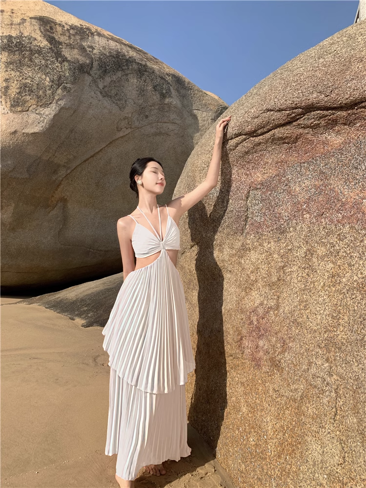 Woman wearing a white halter neck maxi dress with a cut-out and pleated tiered skirt, standing on a beach next to large rocks.