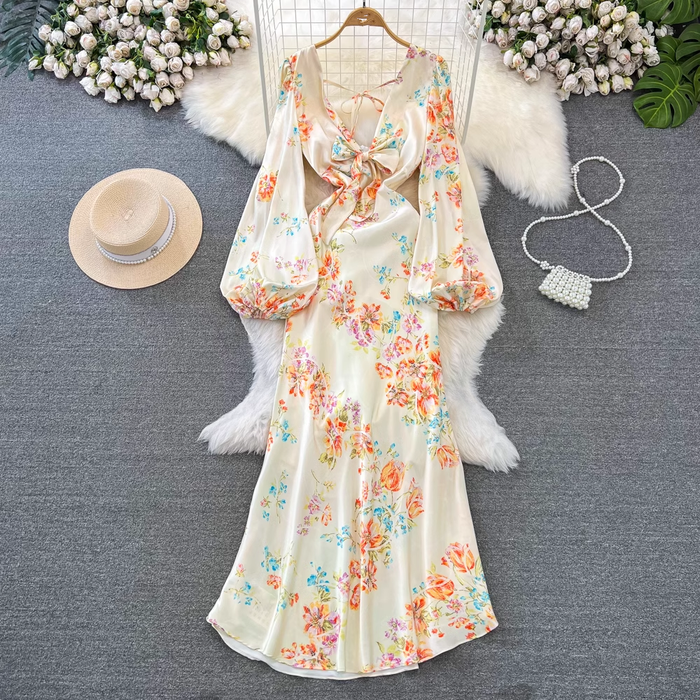 A floral print satin dress with bell sleeves and a bow detail is laid flat on a grey surface with a straw hat and a beaded purse nearby.