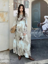 Woman wearing a long-sleeved, tie-dye maxi dress with brown boots and a tan handbag, standing outdoors.