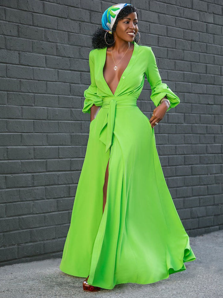 Woman in a lime green maxi dress with a V-neck and high slit, standing against a brick wall.