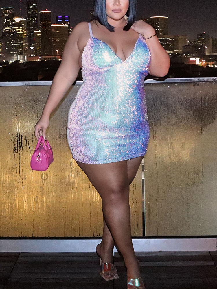 A woman in a shimmering sequined mini dress and holding a pink handbag, standing on a balcony at night with city lights in the background.
