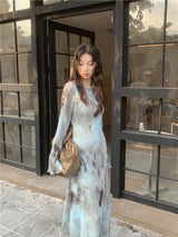 Close-up of a woman wearing a tie-dye maxi dress with a pearl necklace, standing in front of a glass storefront.
