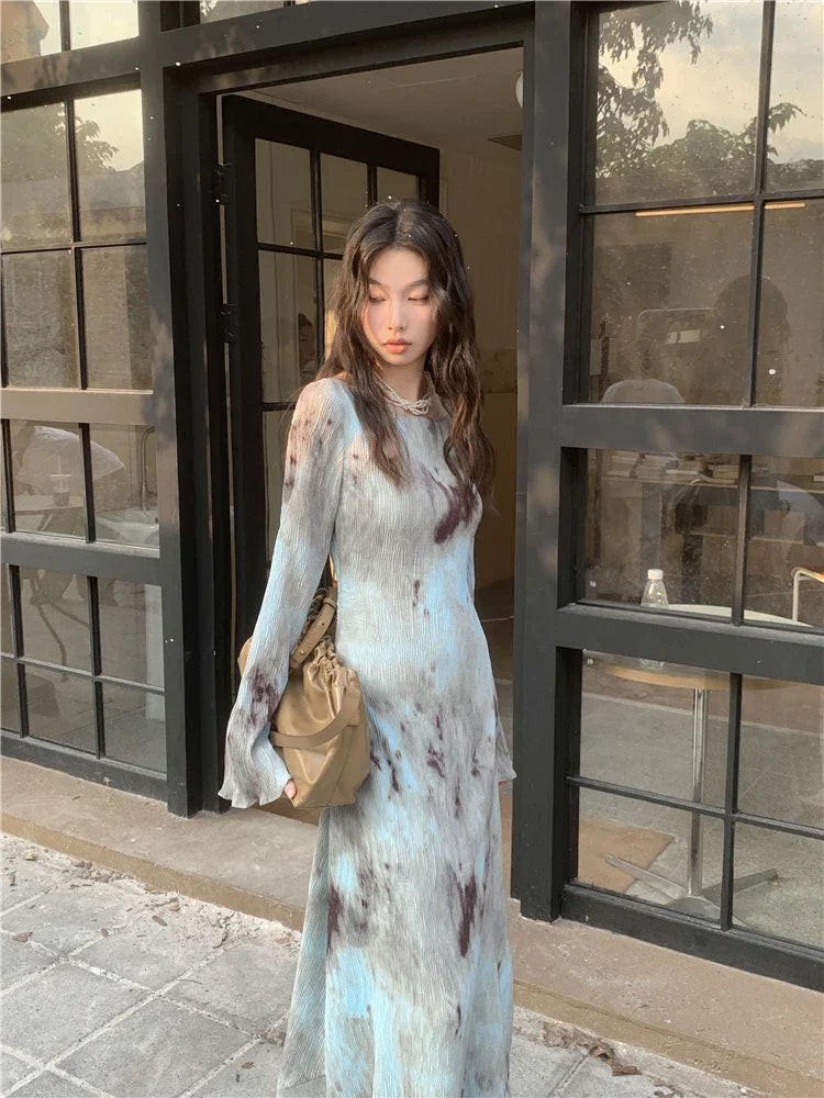 Close-up of a woman wearing a tie-dye maxi dress with a pearl necklace, standing in front of a glass storefront.
