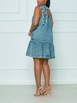 Woman wearing a blue denim A-line dress with tie shoulder straps and a ruffled hem, shown from the back.