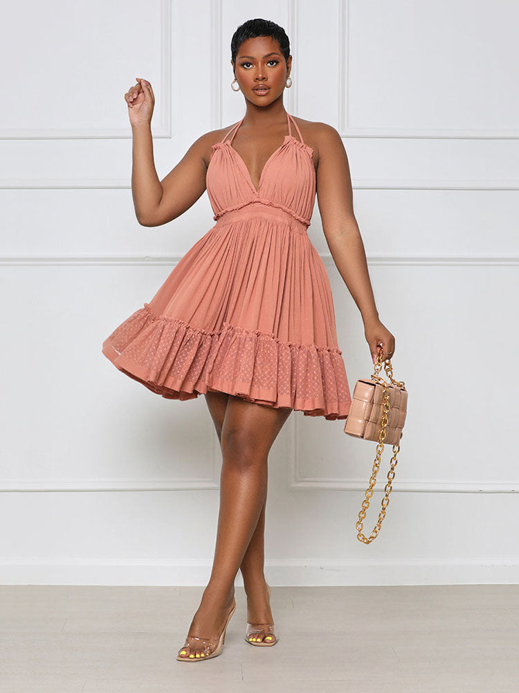 Woman wearing a coral pink halter neck mini dress with a deep V-neckline and tiered ruffled skirt, holding a beige quilted handbag and wearing clear heels.
