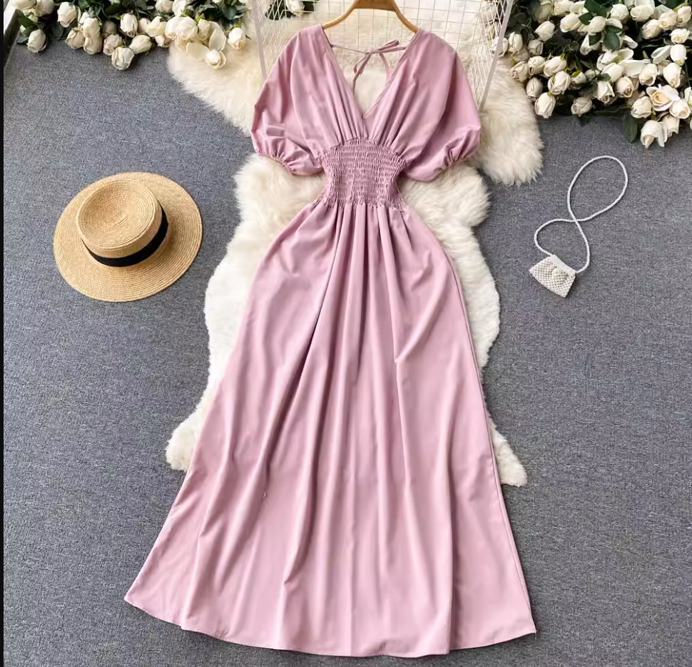 Pink maxi dress with smocked waist and puff sleeves laid flat on a fluffy rug with a straw hat and a small beaded purse