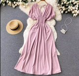 Pink maxi dress with smocked waist and puff sleeves laid flat on a fluffy rug with a straw hat and a small beaded purse