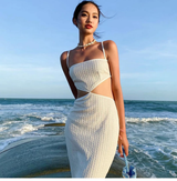Woman wearing a white knit midi dress with cut-out details and a small blue bag, standing by the ocean.