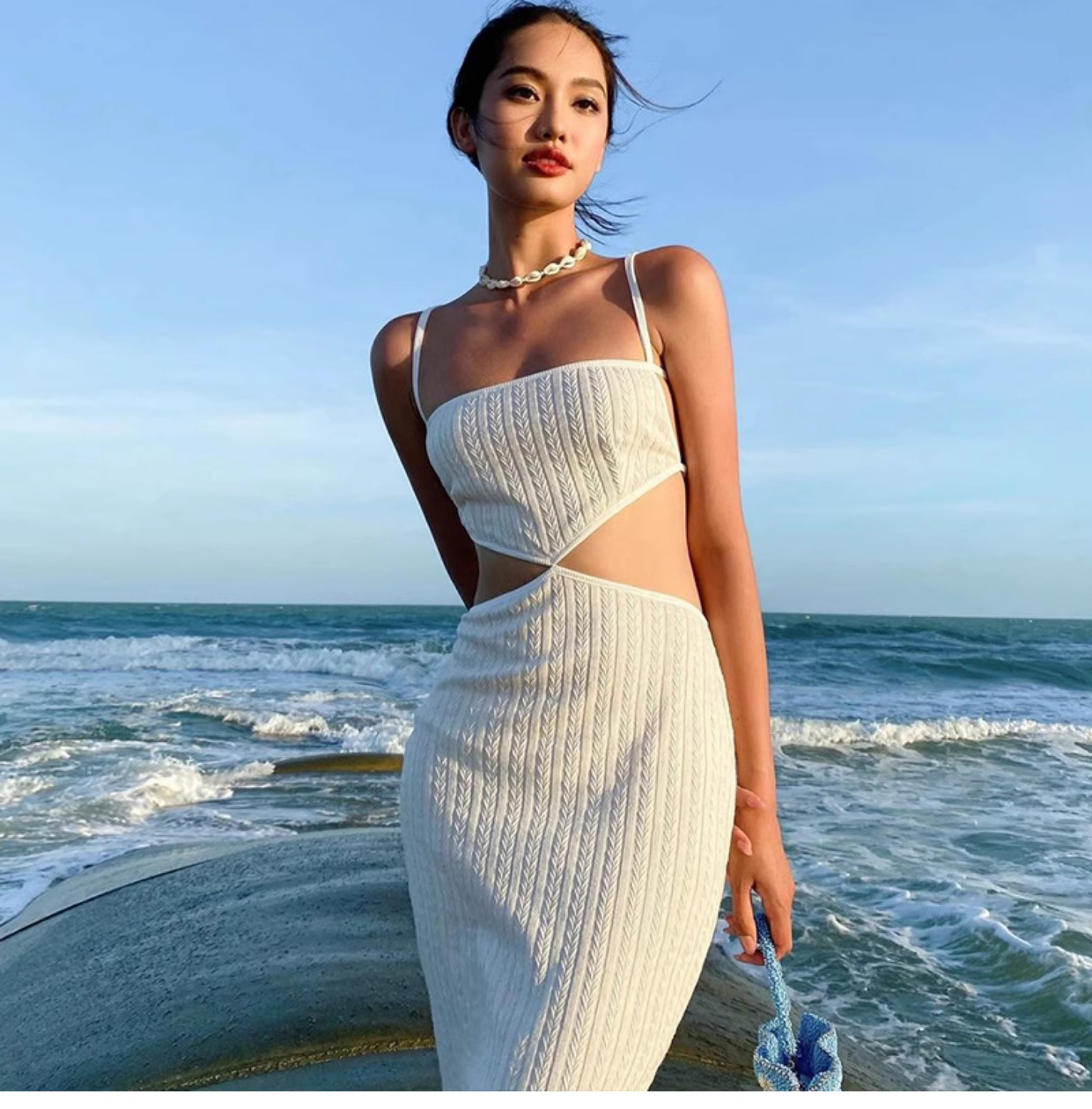 Woman wearing a white knit midi dress with cut-out details and a small blue bag, standing by the ocean.