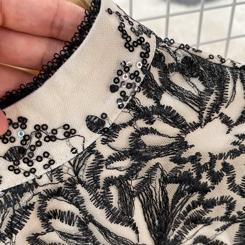 A pair of sparkling silver pointed-toe heels are placed next to a black and white floral embroidered midi dress on a grey surface.