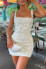 Woman wearing a white sequined mini dress and holding a white clutch bag in a restaurant setting.