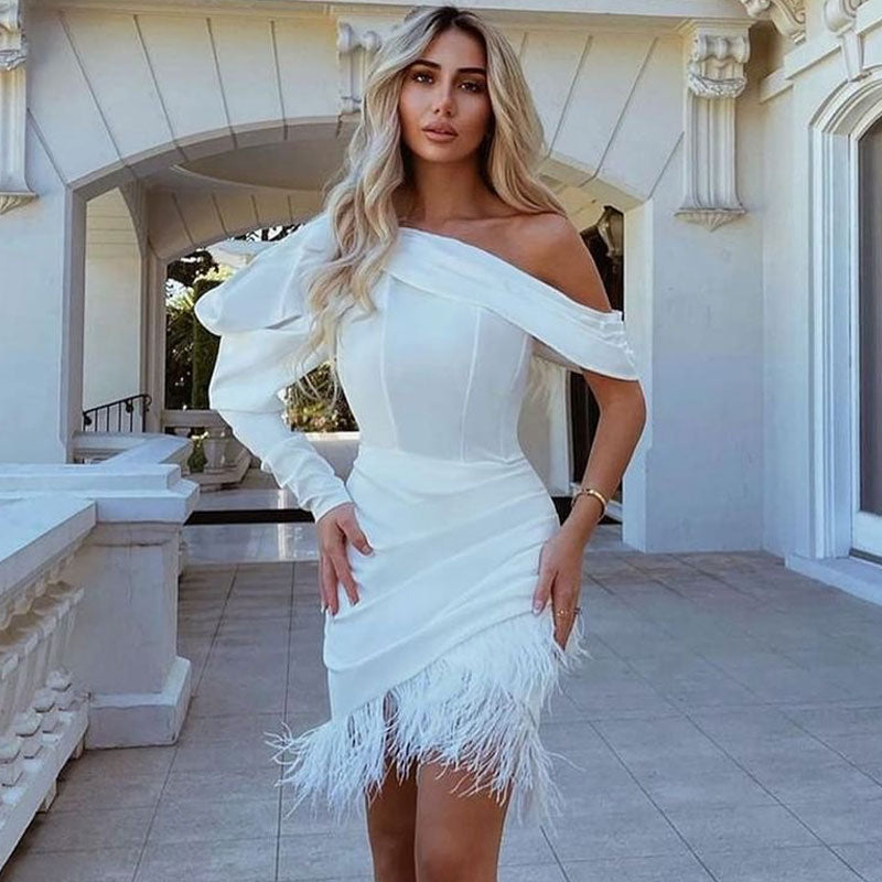 Woman with blonde hair wearing a white asymmetrical mini dress with a feather trim hem and one long sleeve, standing in an outdoor architectural setting.