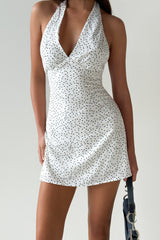 Woman wearing a white halter neck mini dress with black polka dots, holding a dark handbag, against a white background.