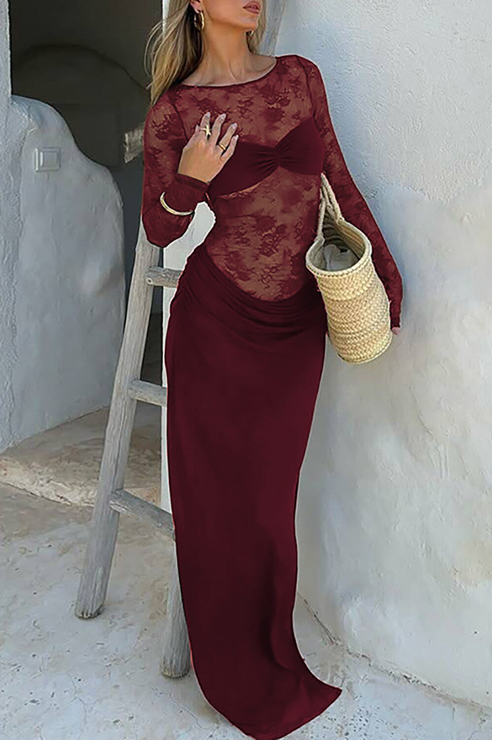 Woman wearing a long burgundy lace dress with a ruched waist and a straw bag.