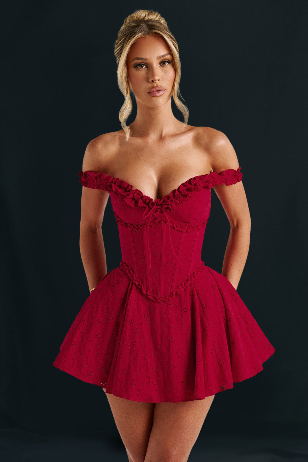 Woman wearing a red off-the-shoulder mini dress with a ruffled neckline and corset bodice, posing against a dark background.