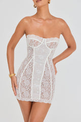 Front close-up of a woman wearing a white strapless lace mini dress with boning details and scalloped hem, accessorized with gold bracelets.