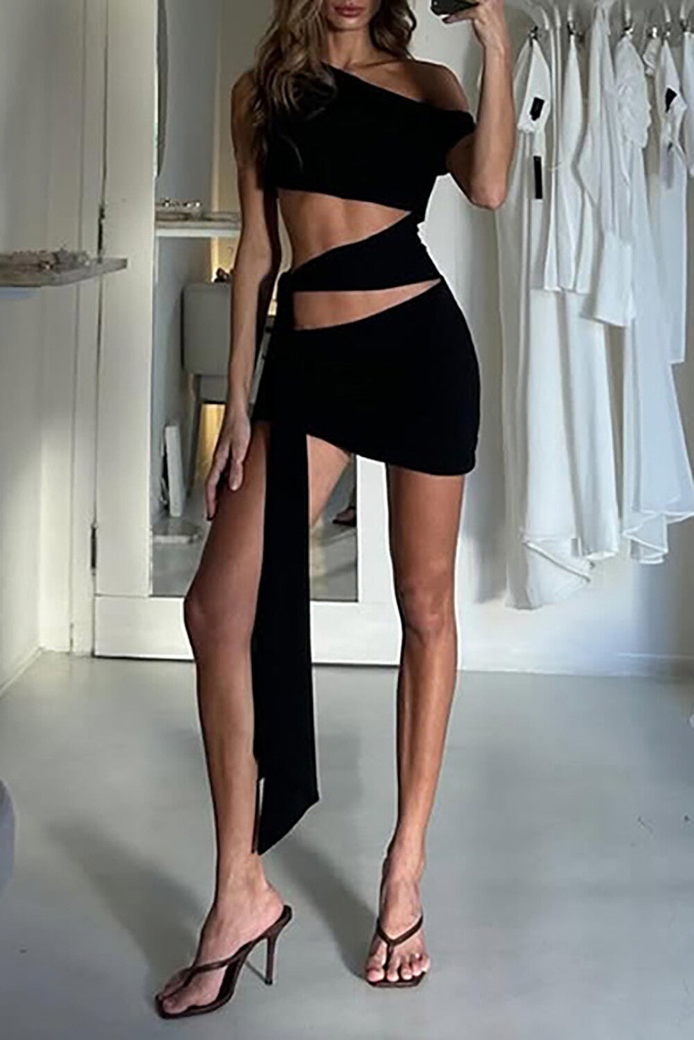 side view of a woman wearing a black one-shoulder asymmetrical cut-out mini dress with a waist tie
