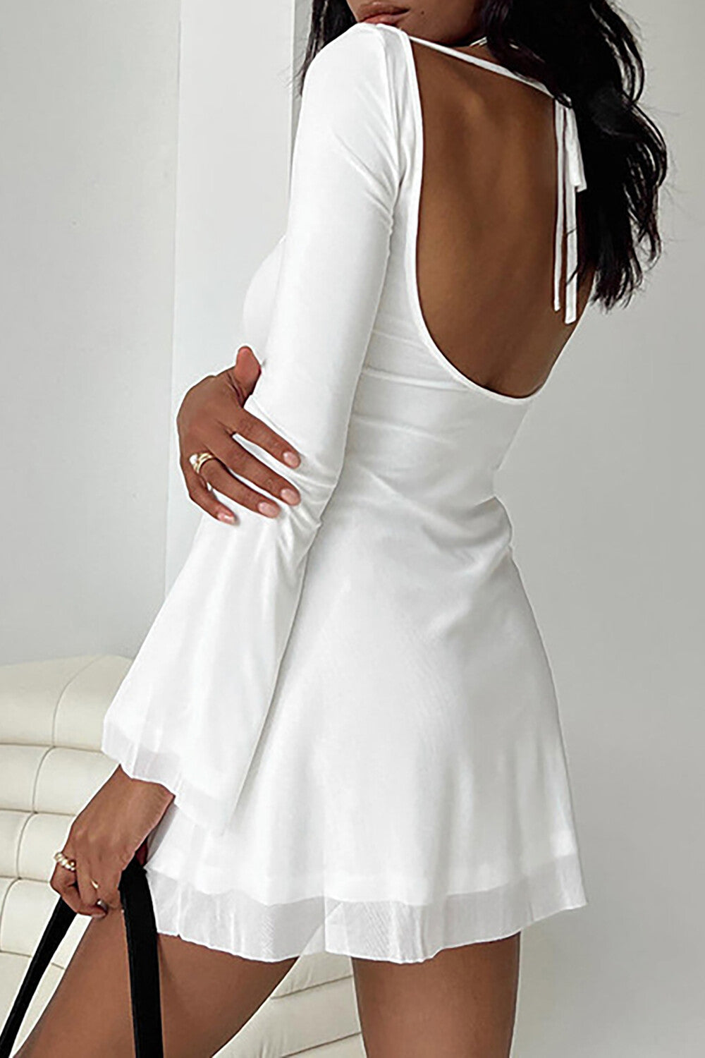 Back view of a woman wearing a white open-back mini dress with tie closure and bell sleeves.