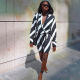 Woman wearing a black and white striped wrap mini dress with long sleeves and a plunging V-neck, paired with black strappy heels and a black shoulder bag, walking on a sidewalk next to a textured wall.