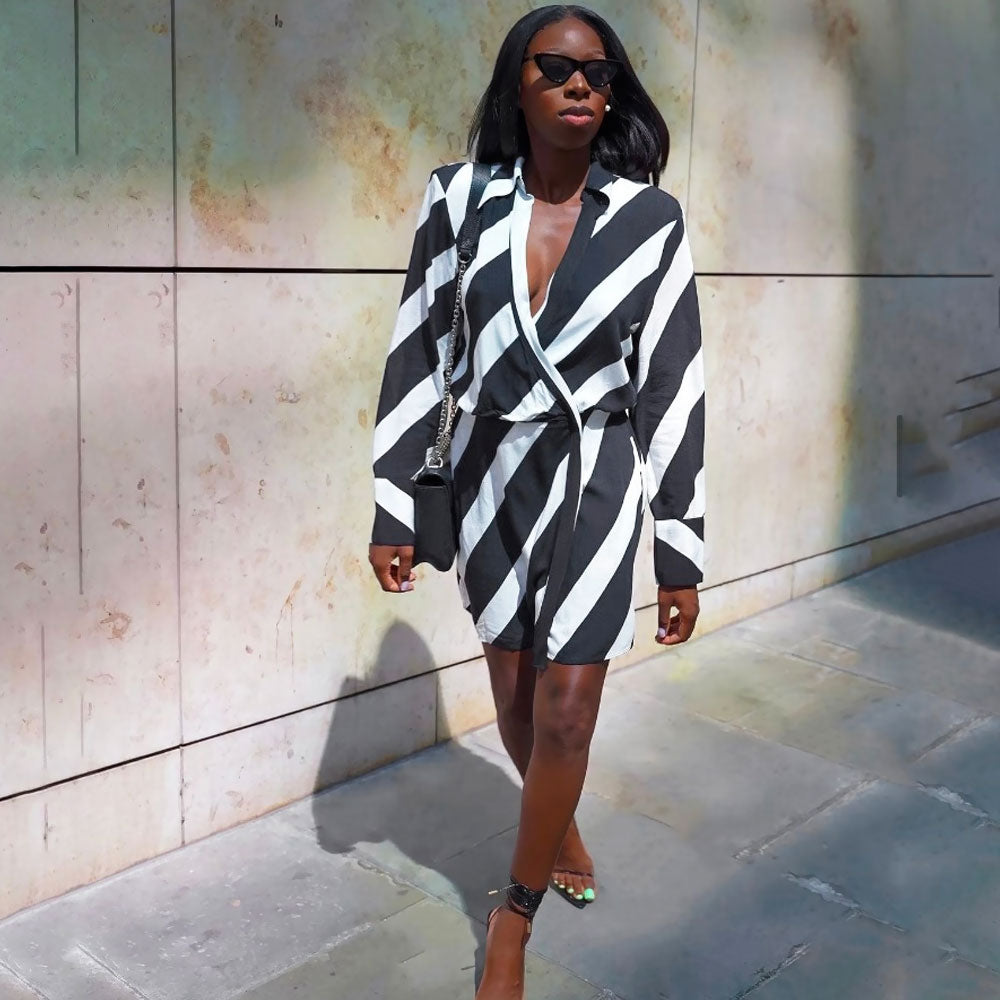 Woman wearing a black and white striped wrap mini dress with long sleeves and a plunging V-neck, paired with black strappy heels and a black shoulder bag, walking on a sidewalk next to a textured wall.
