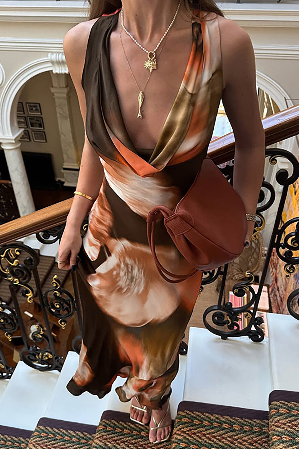 A woman wearing a long, flowing dress with a brown, orange, and white abstract print, a draped neckline, and a brown handbag.