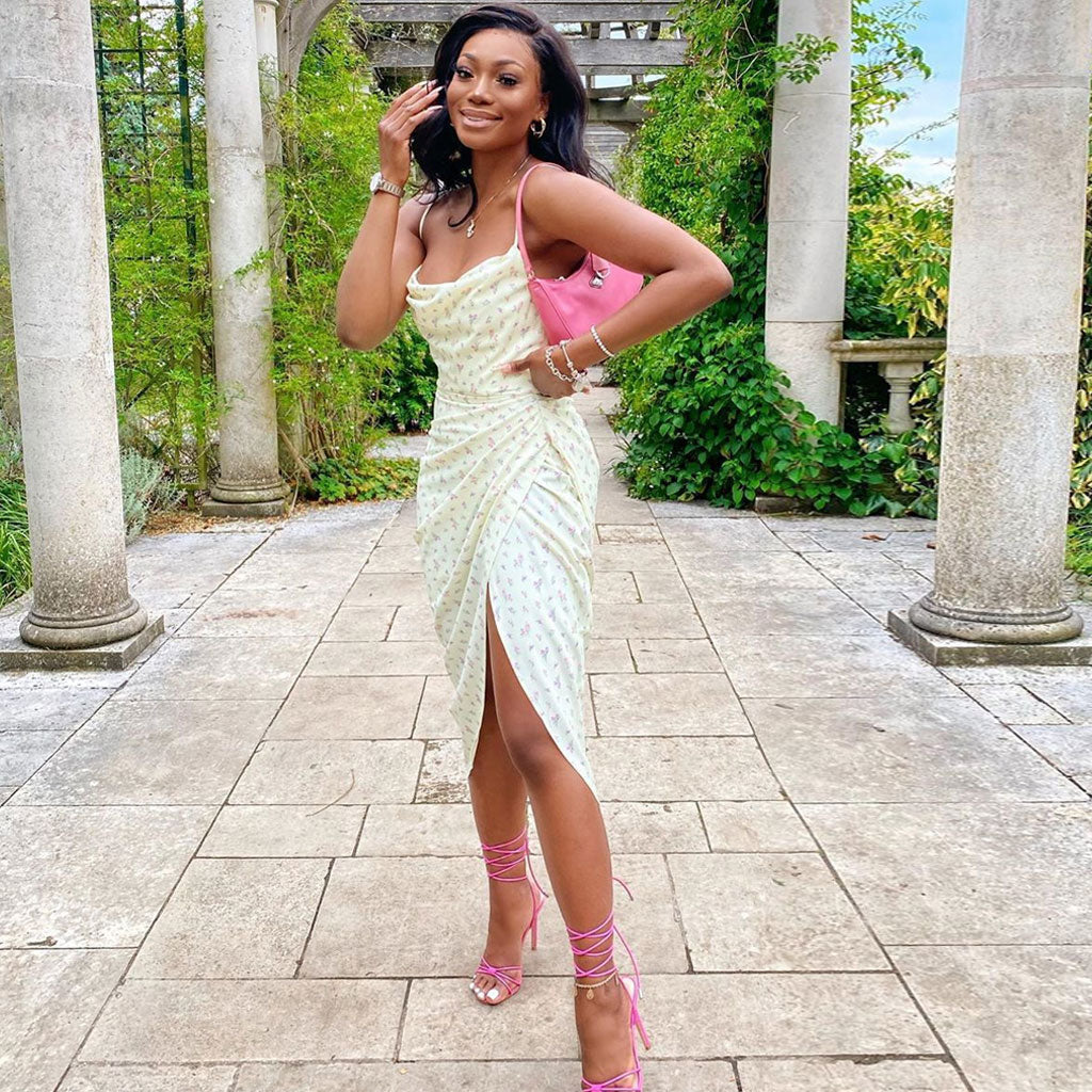 Woman in a yellow floral print satin slip dress with a cowl neck and high slit, posing in a garden with stone columns.