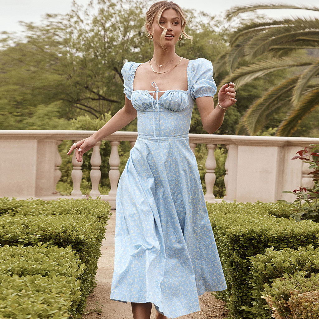 Woman wearing a light blue floral print midi dress with puff sleeves and a square neckline, walking in a garden.